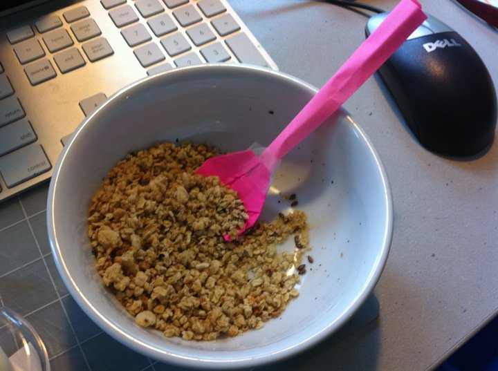 A bowl of granola on a desk with a makeshift spoon made from bright pink sticky notes in the bowl.