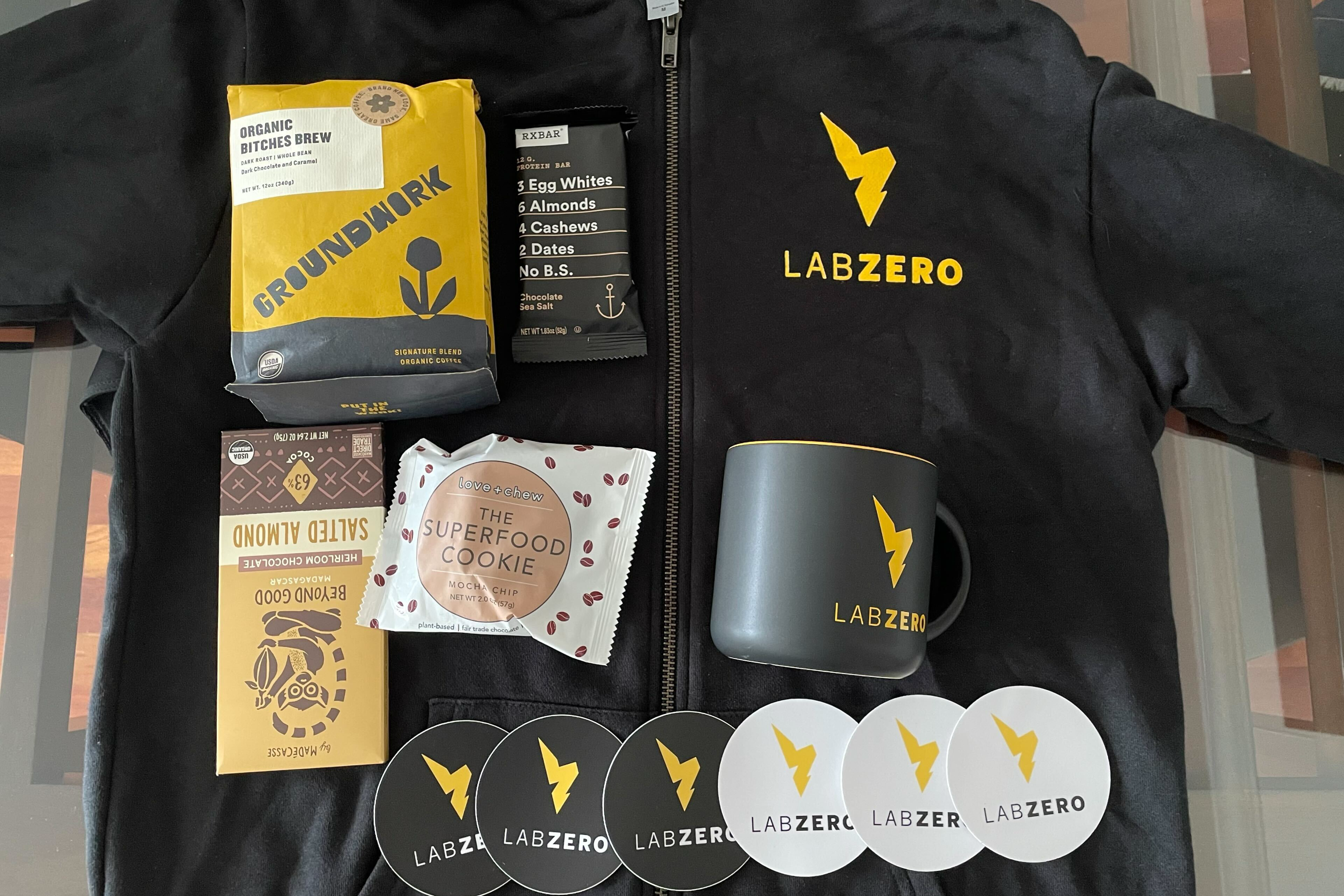 An LZ branded coffee mug, stickers, and a hoodie paired with coffee, chocolate, and other treats. These are part of the welcome kit for new employees.